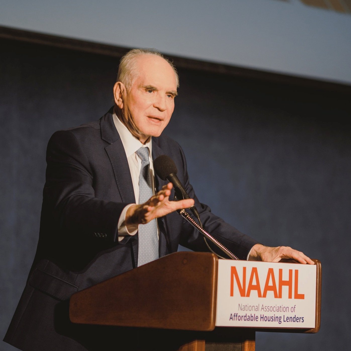 A man in a suit speaking at a podium with a microphone, displaying a sign that reads 'NAAHL National Association of Affordable Housing Lenders.'