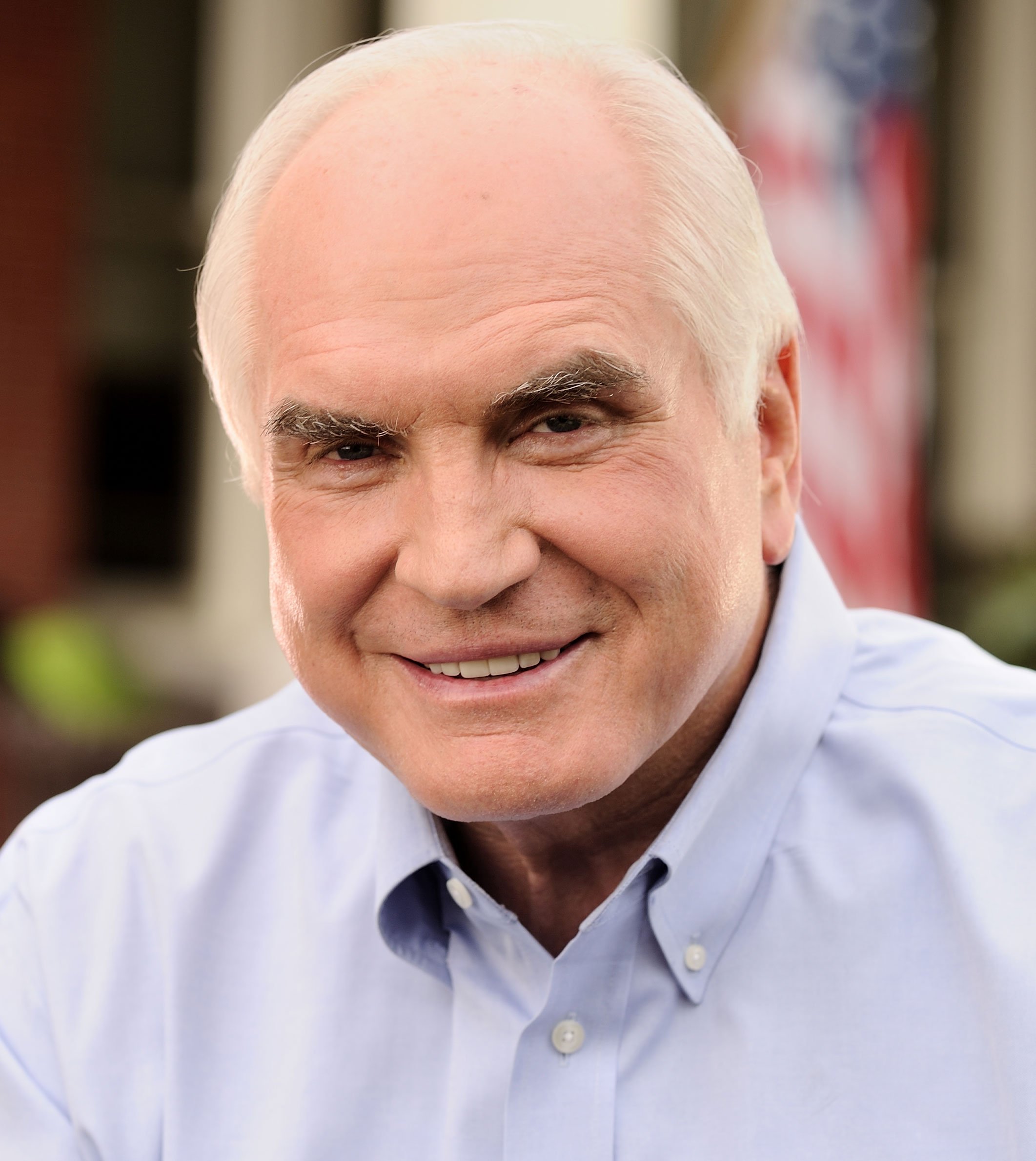 Elderly man smiling in a blue collared shirt with American flag in background.